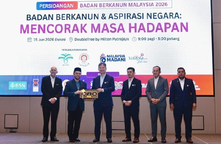 mof kewangan - Wilayah.com.my Six men in suits stand on a stage as one hands a wooden trophy to another, with a large screen behind them displaying an event banner in Malay.