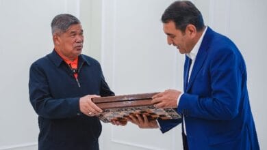 mohamad saba fergana - Wilayah.com.my Two men exchange a wooden-framed award indoors; one in a blue blazer presents it to a man in a dark jacket with an orange shirt.