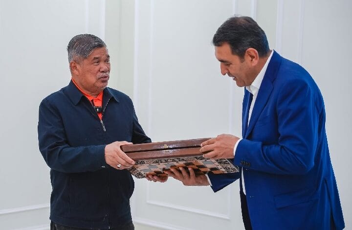 Two men exchange a wooden-framed award indoors; one in a blue blazer presents it to a man in a dark jacket with an orange shirt.