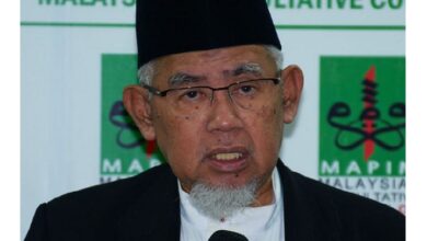 Older man wearing a black cap and glasses speaks into a handheld microphone at a formal event, with a green-and-white backdrop behind him.