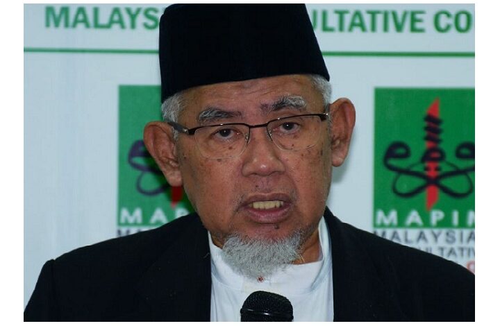 Older man wearing a black cap and glasses speaks into a handheld microphone at a formal event, with a green-and-white backdrop behind him.