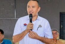 mohd jafni md shukor - Wilayah.com.my Man in a light blue shirt holds a microphone and speaks at an indoor event, with two men seated in the background.