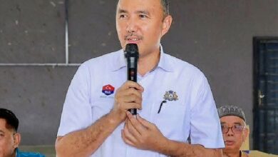 mohd jafni md shukor - Wilayah.com.my Man in a light blue shirt holds a microphone and speaks at an indoor event, with two men seated in the background.