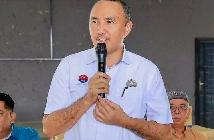 mohd jafni md shukor - Wilayah.com.my Man in a light blue shirt holds a microphone and speaks at an indoor event, with two men seated in the background.
