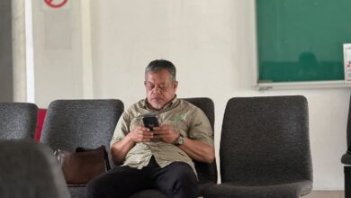 mohd khaidir ahmad - Wilayah.com.my Middle-aged man in a khaki shirt sits in a row of gray chairs, focused on his smartphone in a waiting area.