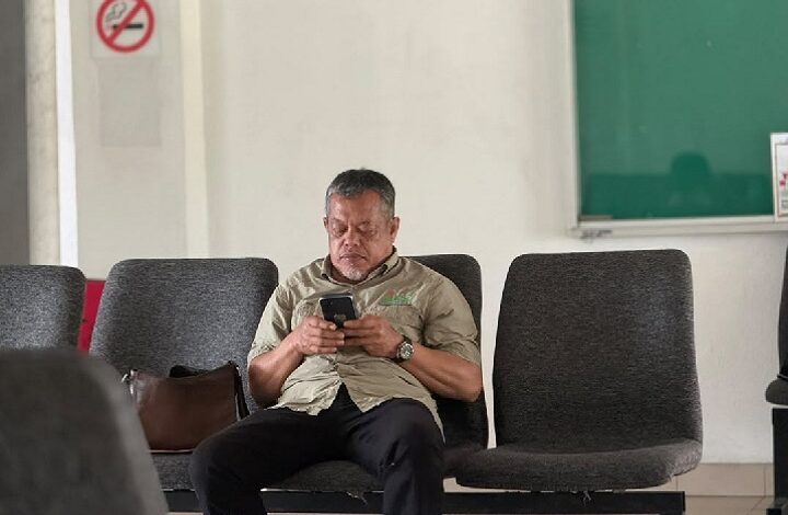 mohd khaidir ahmad - Wilayah.com.my Middle-aged man in a khaki shirt sits in a row of gray chairs, focused on his smartphone in a waiting area.