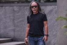 Man with long gray hair and sunglasses standing outdoors in a black T-shirt and blue jeans, facing the camera.