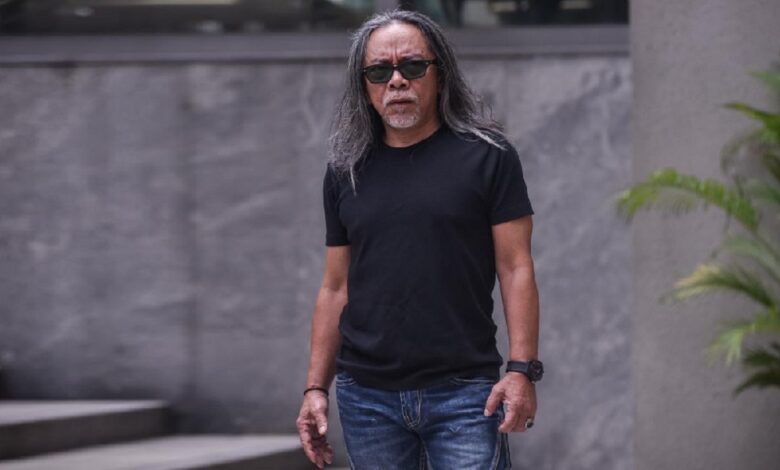 Man with long gray hair and sunglasses standing outdoors in a black T-shirt and blue jeans, facing the camera.