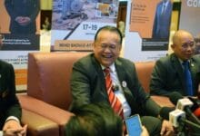 nanta - Wilayah.com.my Smiling man in a dark suit and red striped tie sits on a brown couch at a press conference, with banners behind him and reporters' microphones in front.