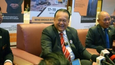 nanta - Wilayah.com.my Smiling man in a dark suit and red striped tie sits on a brown couch at a press conference, with banners behind him and reporters' microphones in front.