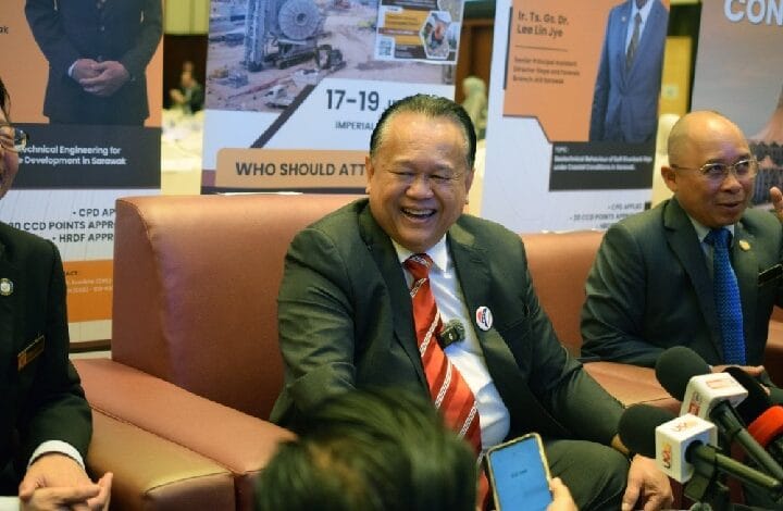 nanta - Wilayah.com.my Smiling man in a dark suit and red striped tie sits on a brown couch at a press conference, with banners behind him and reporters' microphones in front.