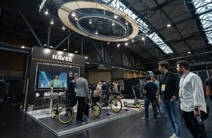 navee - Wilayah.com.my Navée booth at a trade show displaying bicycles with yellow rims; attendees chat and explore the exhibit under a circular overhead light structure.