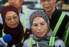 Woman in a patterned hijab speaks to reporters, microphone visible in the foreground; safety vests around her.