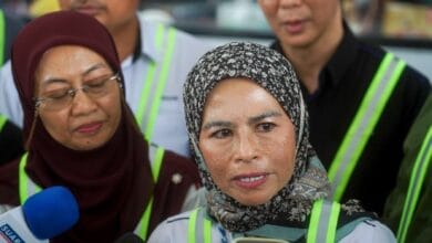 Woman in a patterned hijab speaks to reporters, microphone visible in the foreground; safety vests around her.