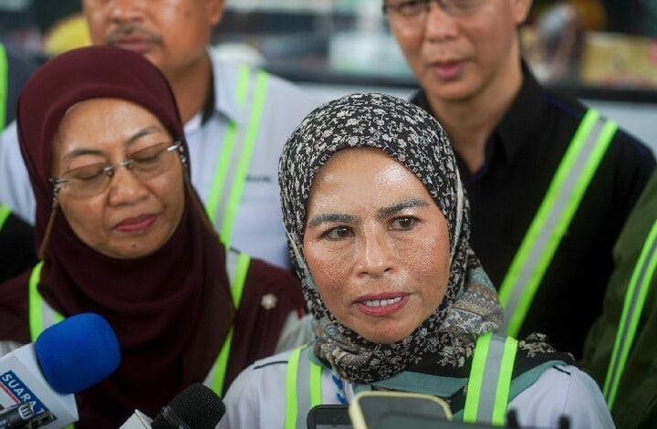 Woman in a patterned hijab speaks to reporters, microphone visible in the foreground; safety vests around her.