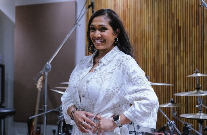 Smiling woman in a white patterned blouse stands in a recording studio with drums and microphone stands in the background.