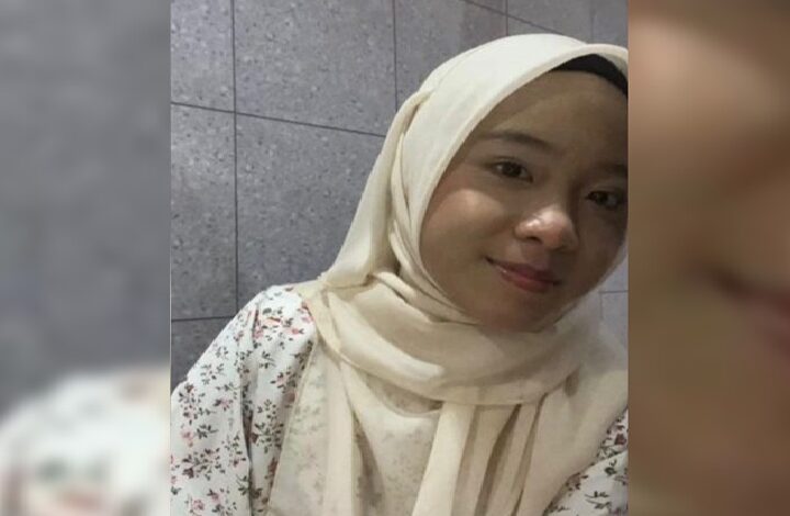 Young woman wearing a cream hijab and floral blouse smiles at the camera against a tiled wall.