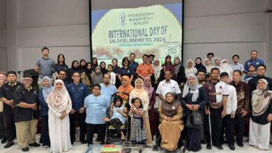 Large diverse group posing indoors for an International Day of Deafblindness 2026 celebration, with a banner in the background and a cake on a table in front.