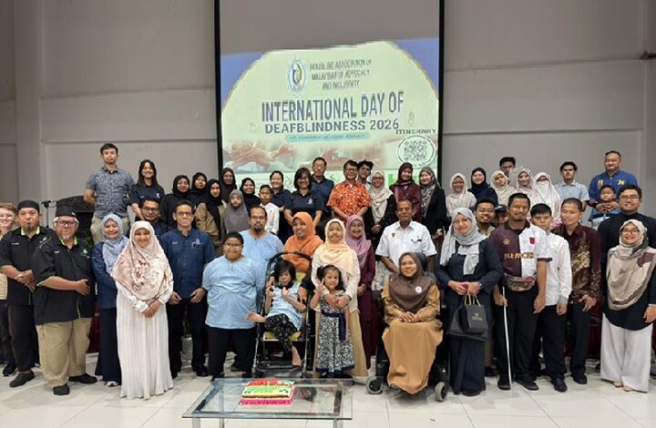 Large diverse group posing indoors for an International Day of Deafblindness 2026 celebration, with a banner in the background and a cake on a table in front.