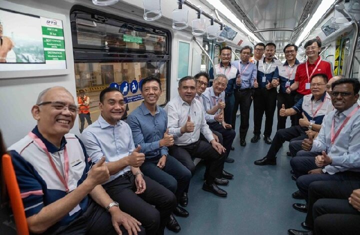 Group of smiling men in business attire seated and standing along both sides of a subway car, giving thumbs up for the camera.