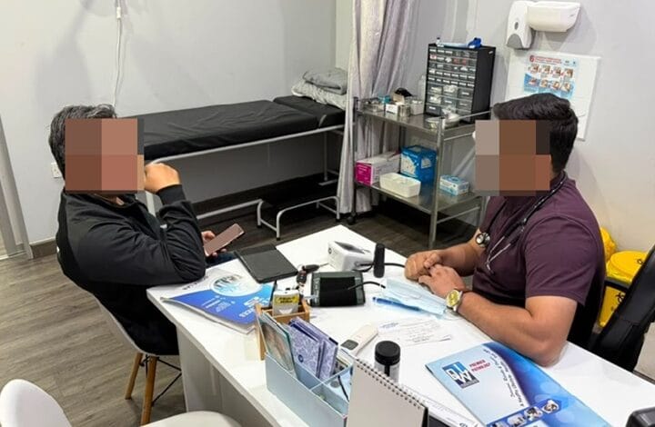 Two people sit across a white desk in a clinic; patient on the left uses a phone while clinician on the right talks.