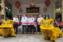 Group of men in white shirts and red ribbons perform a ribbon-cutting ceremony with two yellow dragon/lion dance costumes in front of a temple entrance.