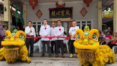 Group of men in white shirts and red ribbons perform a ribbon-cutting ceremony with two yellow dragon/lion dance costumes in front of a temple entrance.