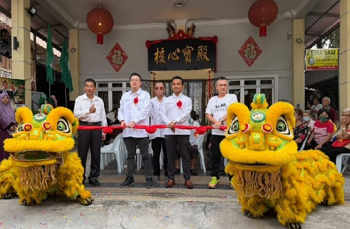 Group of men in white shirts and red ribbons perform a ribbon-cutting ceremony with two yellow dragon/lion dance costumes in front of a temple entrance.