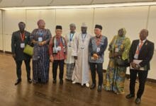 Eight people in formal and traditional attire with conference badges stand in a line inside a conference hall, posing for a group photo.