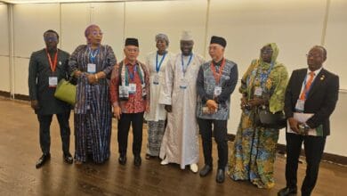 Eight people in formal and traditional attire with conference badges stand in a line inside a conference hall, posing for a group photo.