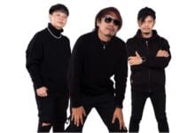 Three men dressed in black outfits pose together against a white background; one wears sunglasses and a necklace while the others stand with folded arms or hands on knees.
