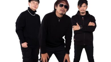 Three men dressed in black outfits pose together against a white background; one wears sunglasses and a necklace while the others stand with folded arms or hands on knees.