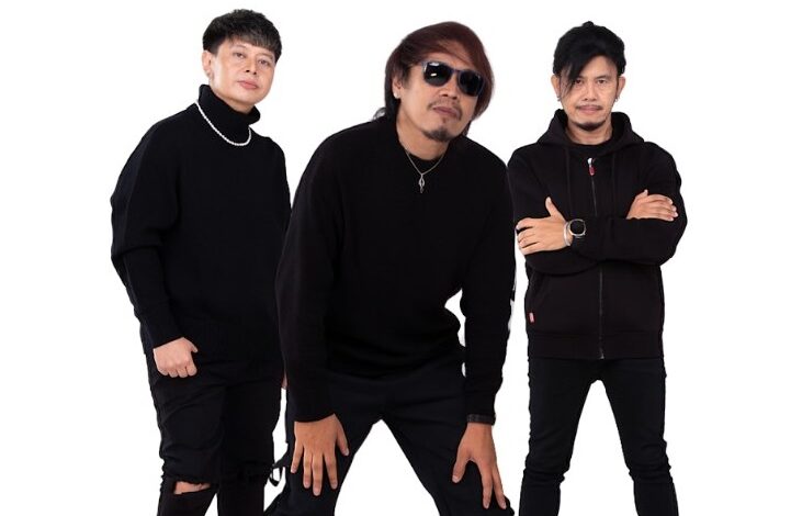 Three men dressed in black outfits pose together against a white background; one wears sunglasses and a necklace while the others stand with folded arms or hands on knees.
