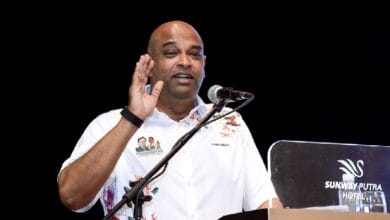 ramanan 2 - Wilayah.com.my Male speaker raises hand while addressing an audience at a podium; wearing a white floral shirt, microphone in front, Sunway Putra Hotel backdrop