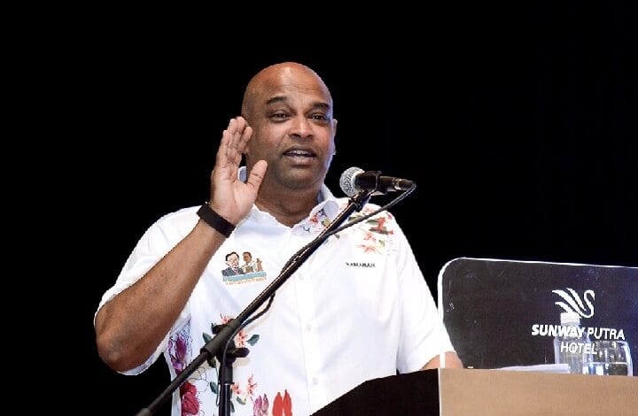 ramanan 2 - Wilayah.com.my Male speaker raises hand while addressing an audience at a podium; wearing a white floral shirt, microphone in front, Sunway Putra Hotel backdrop