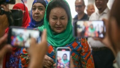rosmah mansor - Wilayah.com.my Woman wearing a green headscarf and colorful dress being photographed by multiple people holding smartphones, with a crowd in the background.