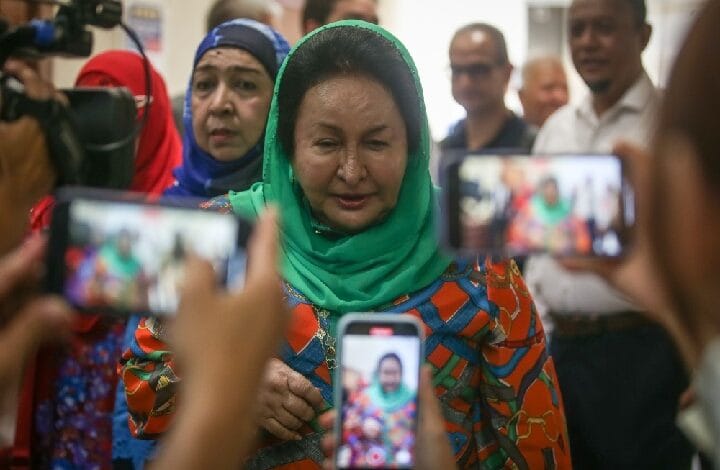 rosmah mansor - Wilayah.com.my Woman wearing a green headscarf and colorful dress being photographed by multiple people holding smartphones, with a crowd in the background.