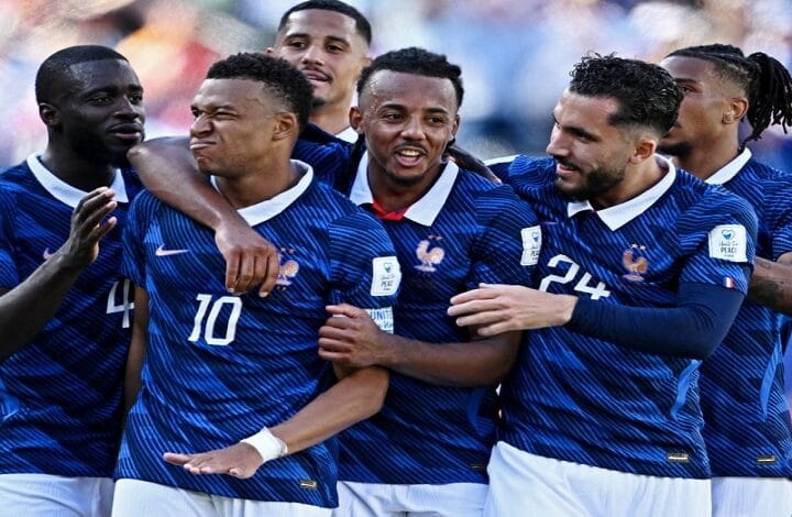 Group of soccer players in blue jerseys celebrate together on the field, arms around each other.”,