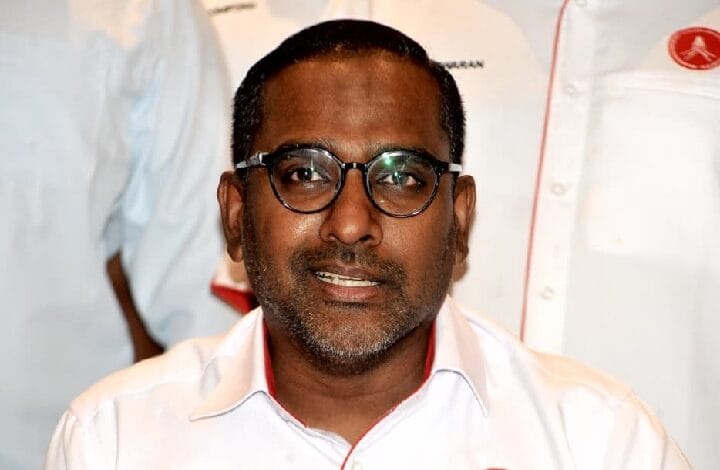 Man with dark hair and round glasses wearing a white shirt with red piping, smiling at the camera.
