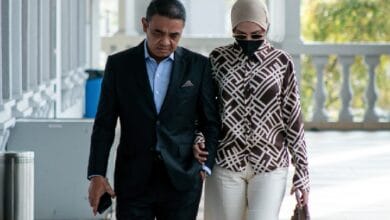 salwani anuar - Wilayah.com.my Man in a black suit walks arm-in-arm with a woman wearing a patterned coat, hijab, mask, and sunglasses outdoors on a sunny day.