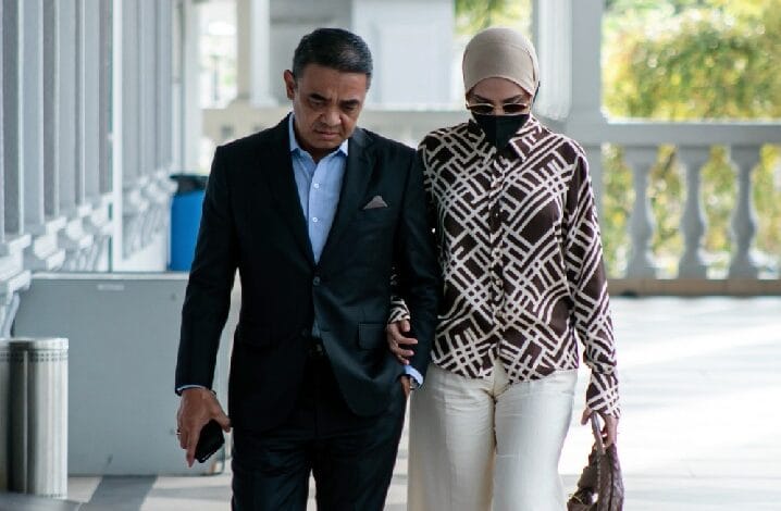 Man in a black suit walks arm-in-arm with a woman wearing a patterned coat, hijab, mask, and sunglasses outdoors on a sunny day.