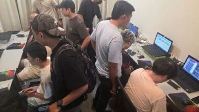 Group of people working at a long table with laptops in a tech workshop setting, some standing and others seated around screens.