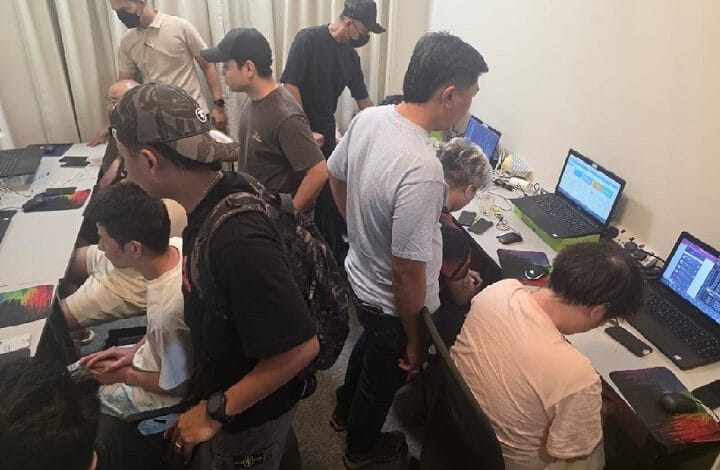 Group of people working at a long table with laptops in a tech workshop setting, some standing and others seated around screens.