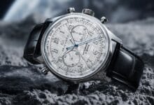 Luxury chronograph wristwatch with a silver dial, three subdials and blue hands on a black leather strap resting on rocky terrain.