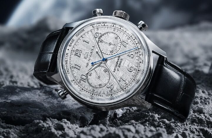 Luxury chronograph wristwatch with a silver dial, three subdials and blue hands on a black leather strap resting on rocky terrain.