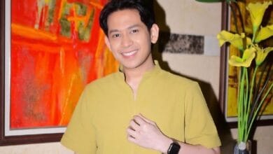 shaffiq erwin - Wilayah.com.my Smiling man with short black hair in a yellow shirt posing indoors near colorful abstract art and yellow flowers.