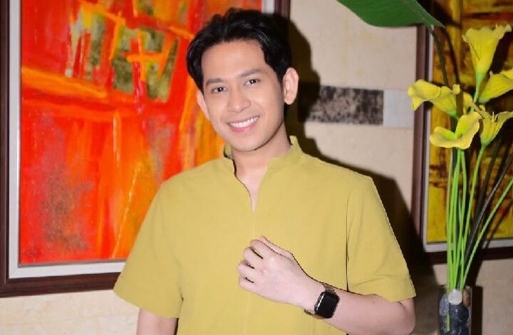 shaffiq erwin - Wilayah.com.my Smiling man with short black hair in a yellow shirt posing indoors near colorful abstract art and yellow flowers.