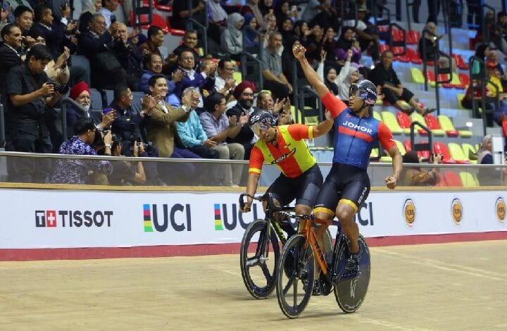 Two cyclists celebrate a victory on a wooden velodrome as the crowd cheers in the stands behind them.