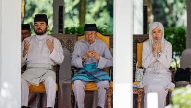 Three people in traditional Malay attire sit in a shaded pavilion, hands raised in prayer.