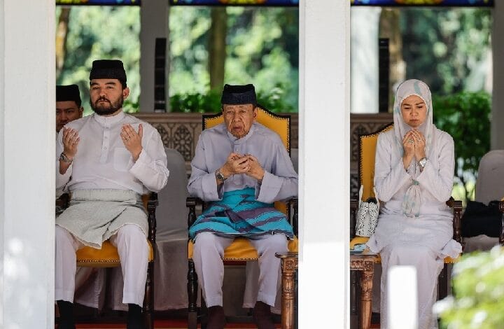 Three people in traditional Malay attire sit in a shaded pavilion, hands raised in prayer.